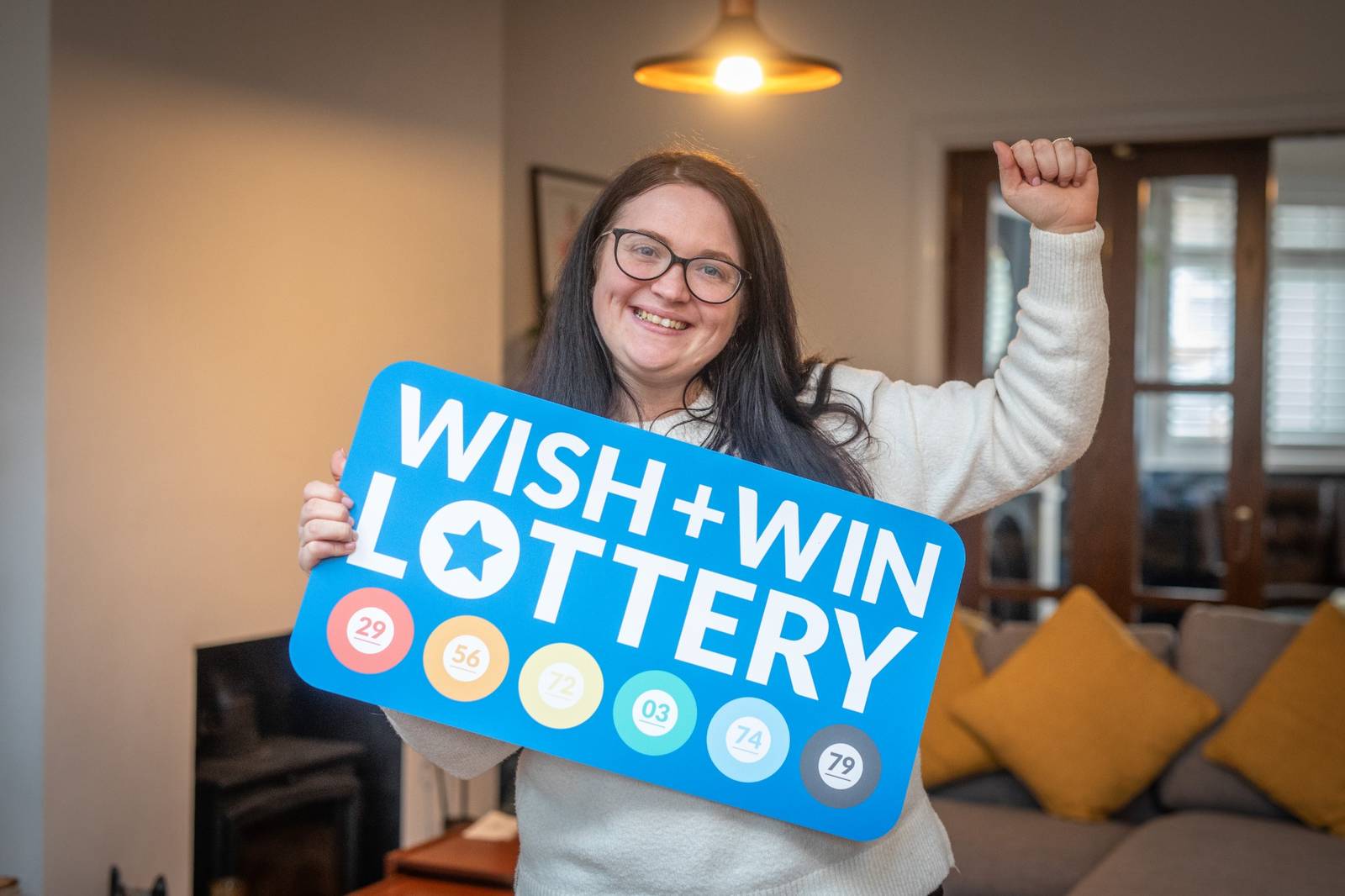 Lottery second prize winner holding a Wish + Win Lottery sign inside her living room